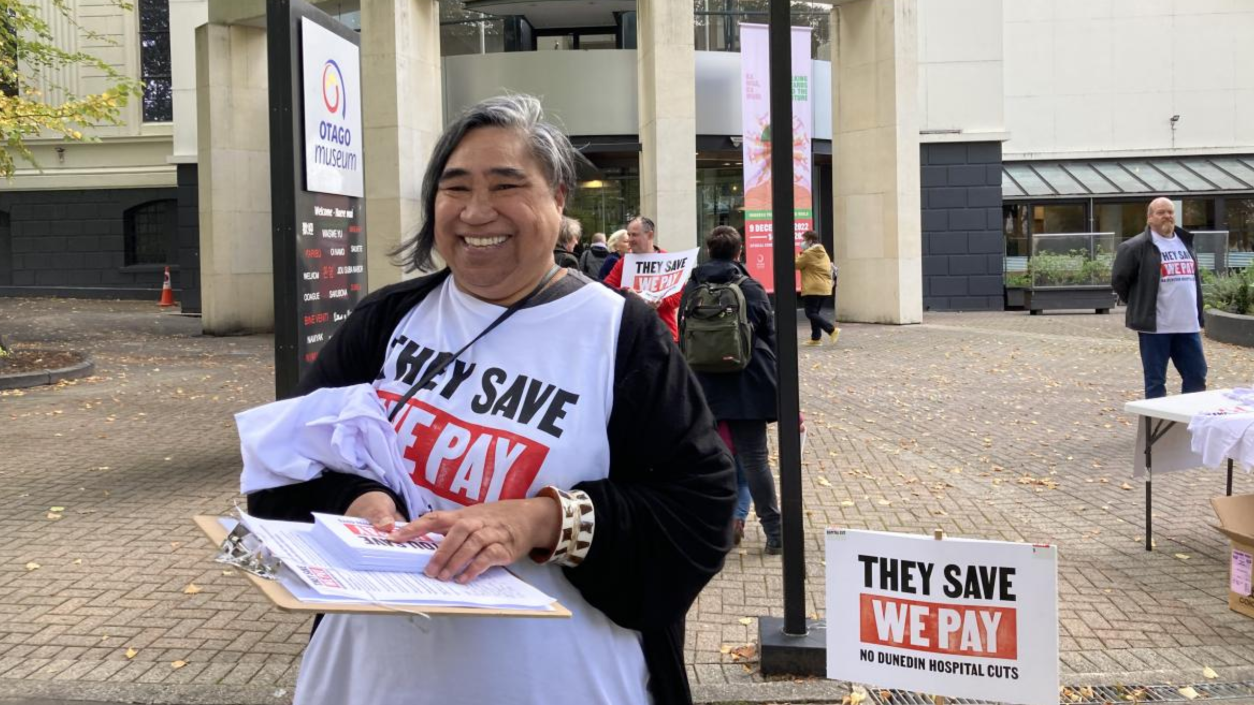 Cr Marie Laufiso is wearing a black cardigan overtop of a white tee-shirt which says "They Save, We Pay". She is holding flyers, and has a purse around her neck. She is standing in front of a sign that also says "They Save, We Pay". She is standing outside of the Otago Museum, several other people are also walking around. 