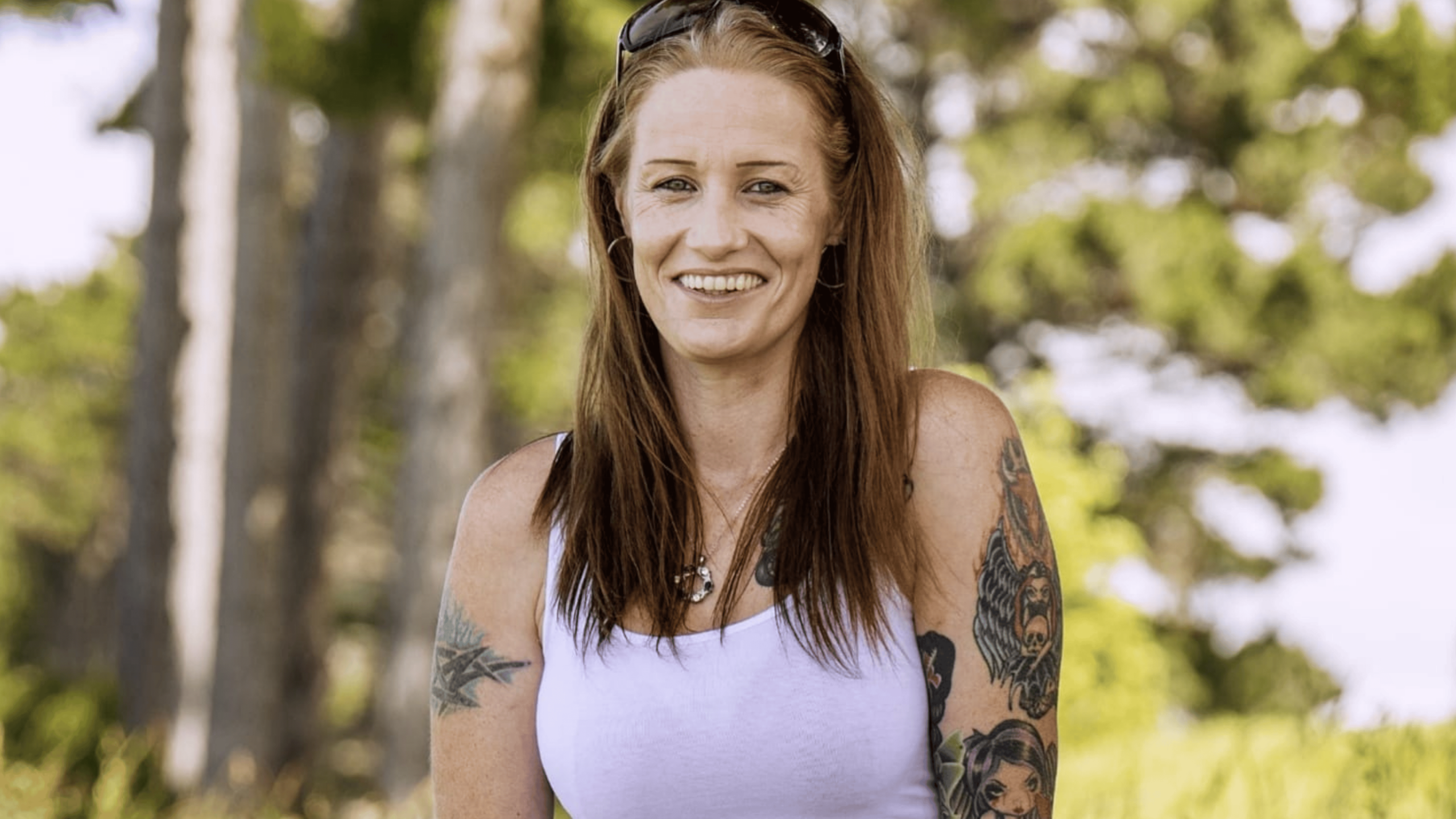 Cyndee Elder sitting in front of a leafy green background wearing a white tank top.