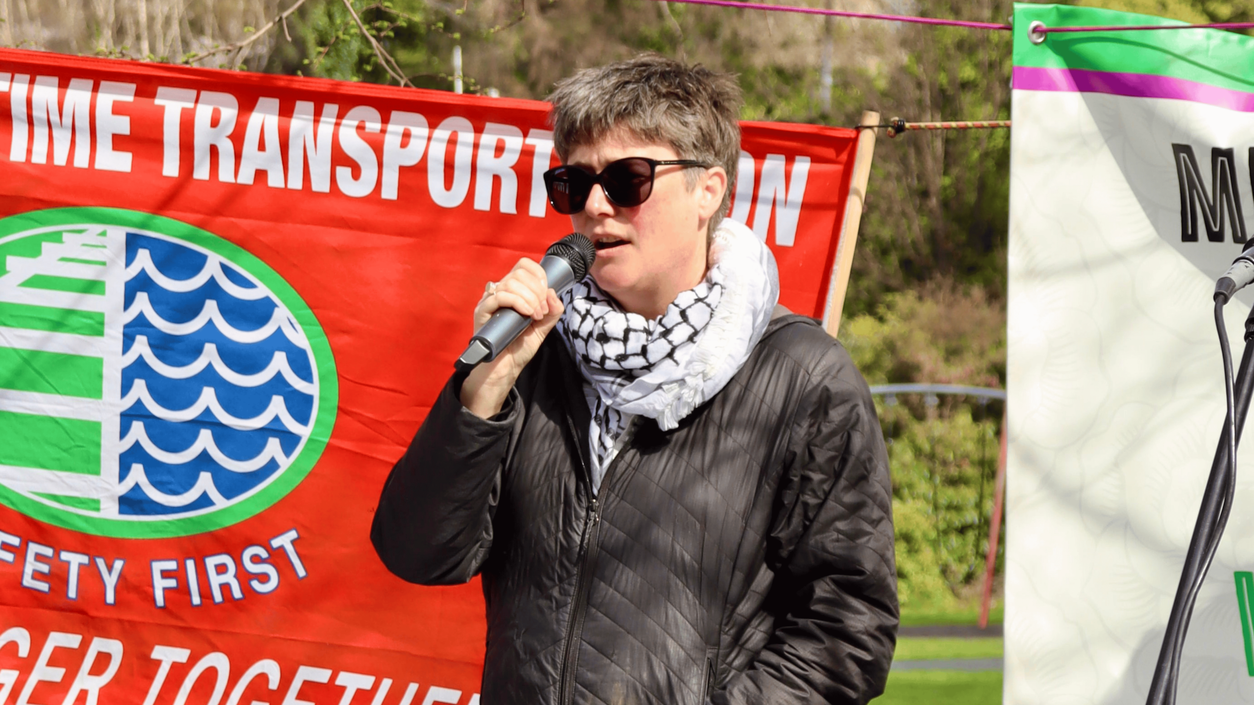 Anna Knight wearing a black puffer jacket, sunglasses, and a keffiyeh holding a microphone. She is standing in front of a red flag for the Rail and Maritime Transport Union